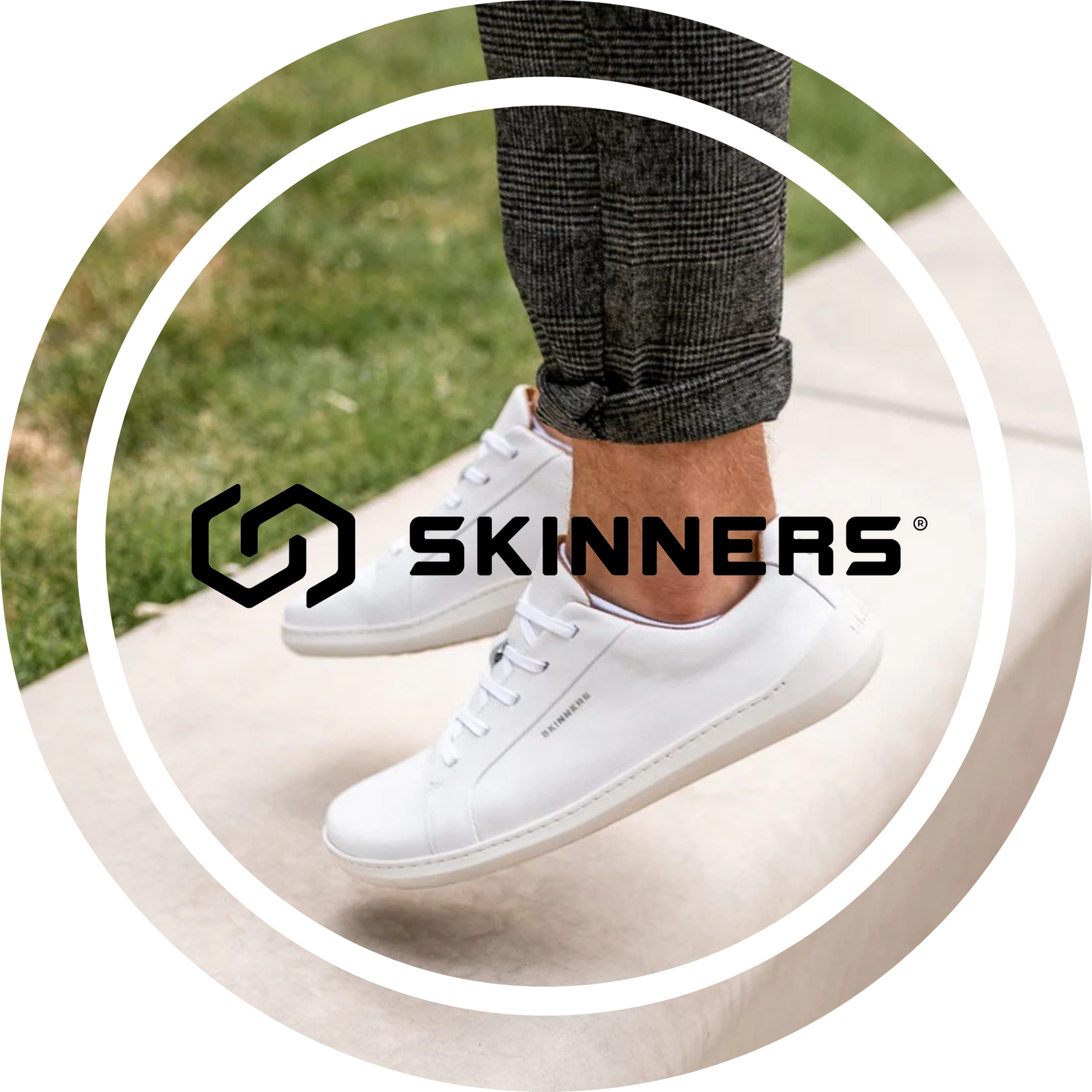 Person med uppvikta grårutiga byxor bär vita minimalistiska sneakers från Skinners, stående på en betongkant med gräs i bakgrunden. Bilden är rund med vit ram och Skinners-loggan i svart centrerad ovanpå.