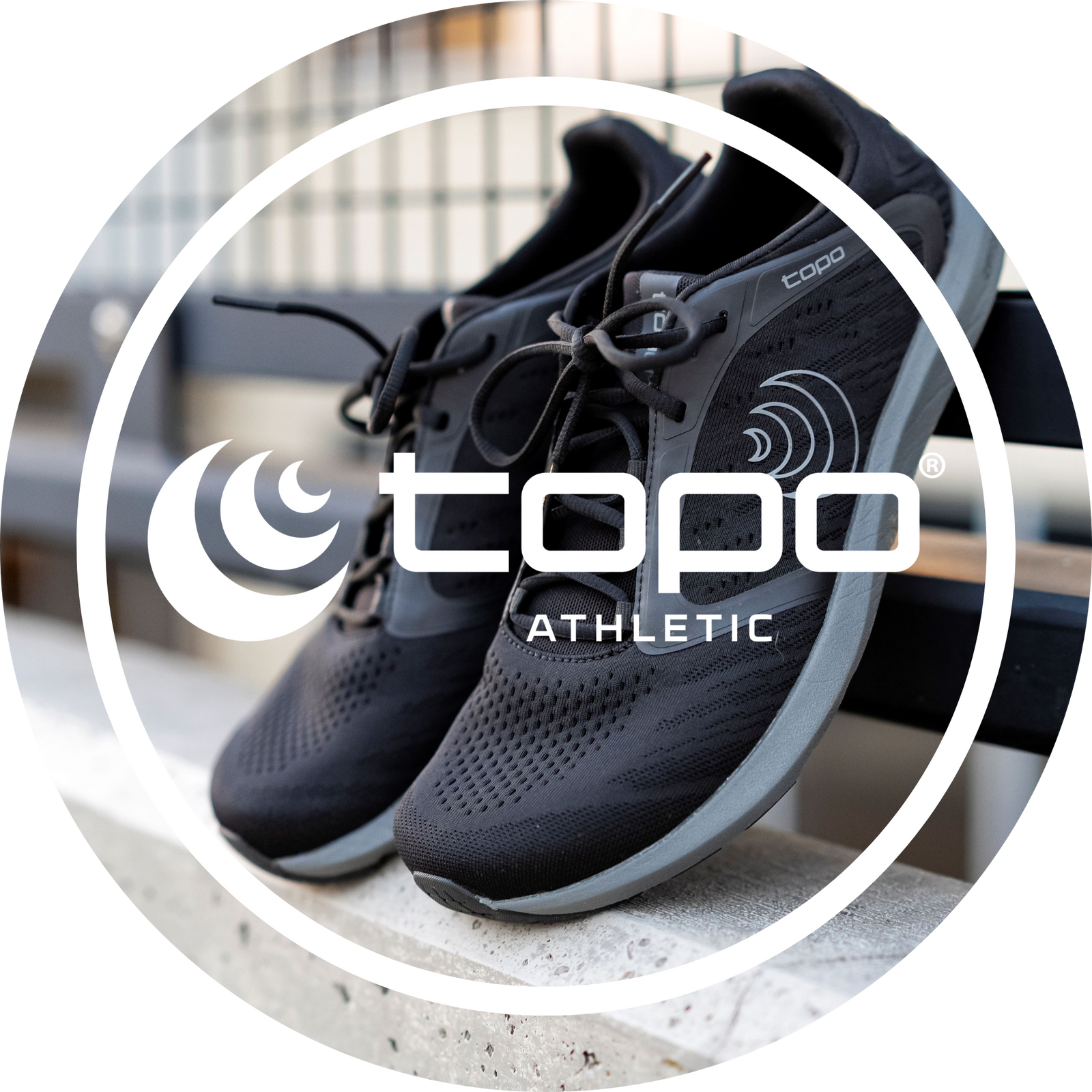 Svart löparsko från Topo Athletic med grå sula, designad för naturlig löpning – produktfoto med logotyp i fokus på en stadsmiljöbänk.