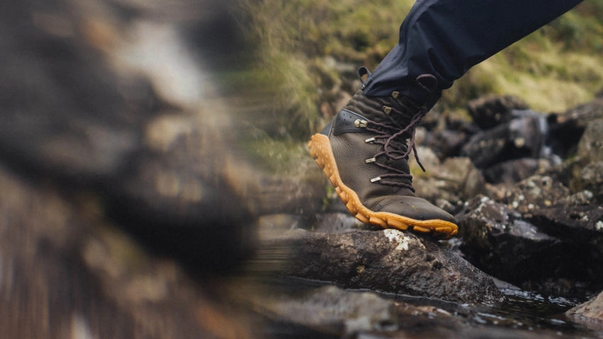 Närbild på en person som korsar ett stenigt vattendrag i ett par Vivobarefoot Tracker Forest ESC-kängor i färgen bracken med ljusbrun sula. Personen bär mörka byxor och syns från knäet och nedåt i ett utomhuslandskap med mossa och stenar.