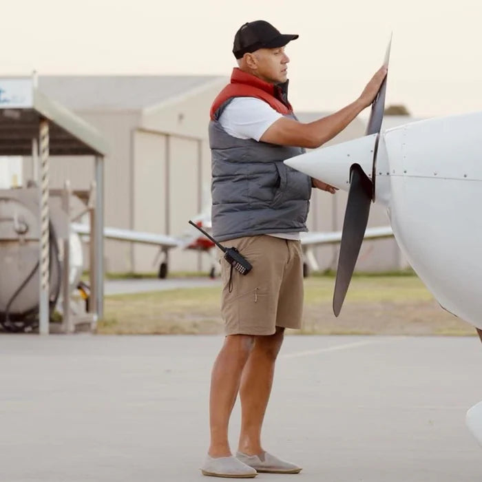 Man i funktionskläder och minimalistiska slip-on skor står vid ett litet flygplan och inspekterar propellern på en flygplats