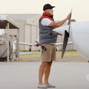 Man i funktionskläder och minimalistiska slip-on skor står vid ett litet flygplan och inspekterar propellern på en flygplats