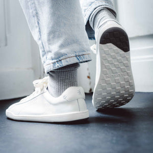 Person med ljusa jeans och gråa strumpor bär vita barfotaskor från Blusun. Fokus på skornas flexibla yttersula med greppmönster och märkets logotyp synlig i hälen.